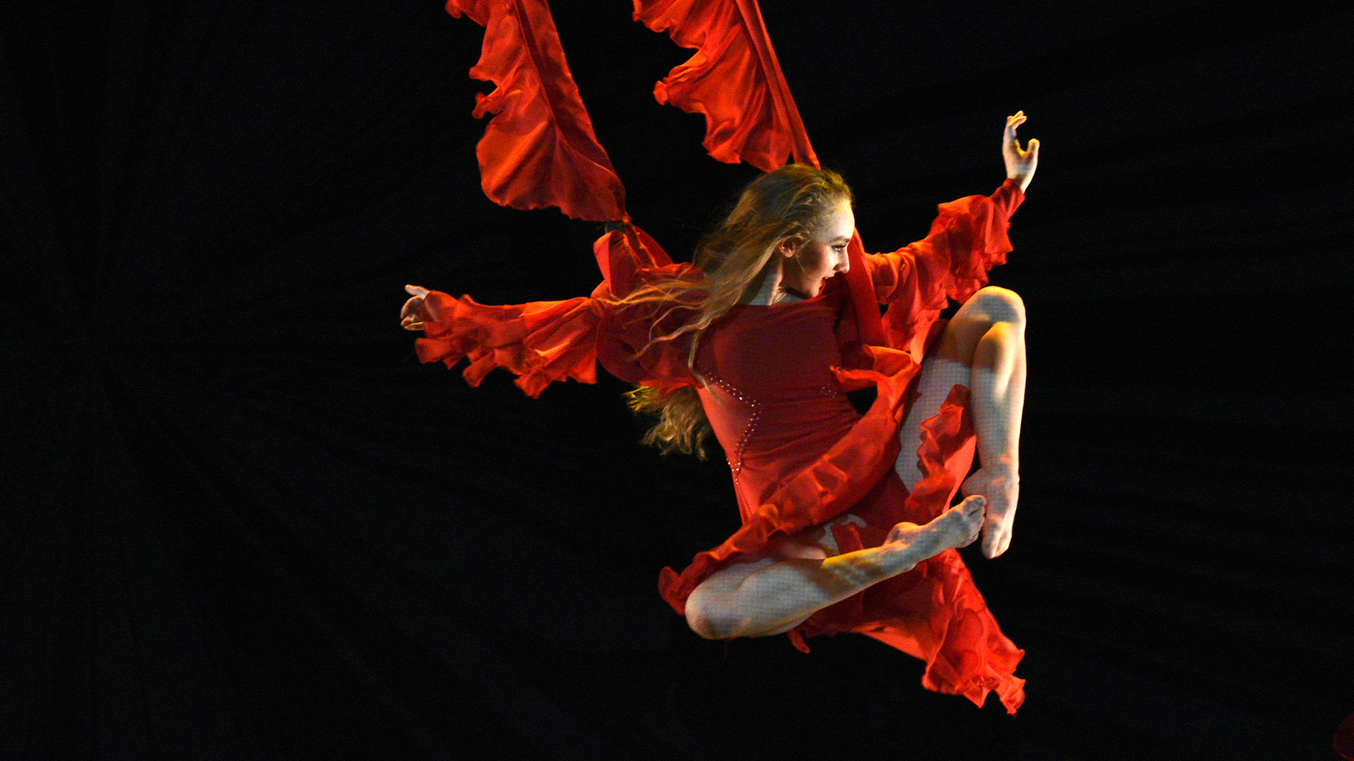 Female dancer wearing a red frilly costume flying in from the left on a trapeze