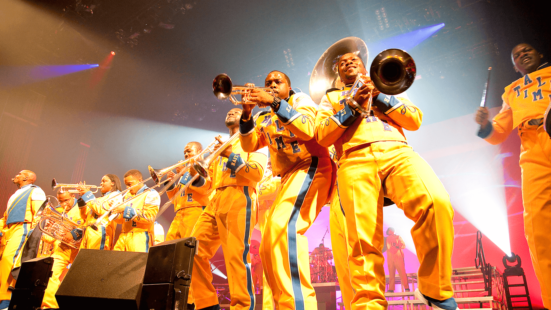 People in yellow drumline suits stand with various brass instruments on stage