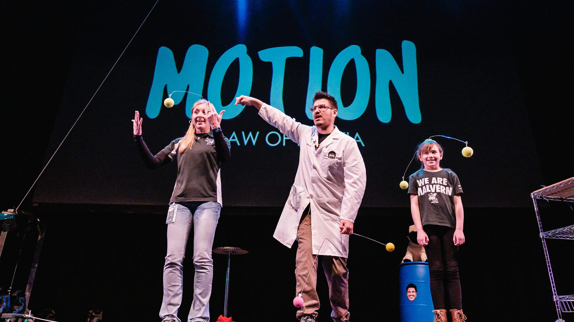 A man in a lab coat and two people on stage with tennis ball floating in the air 