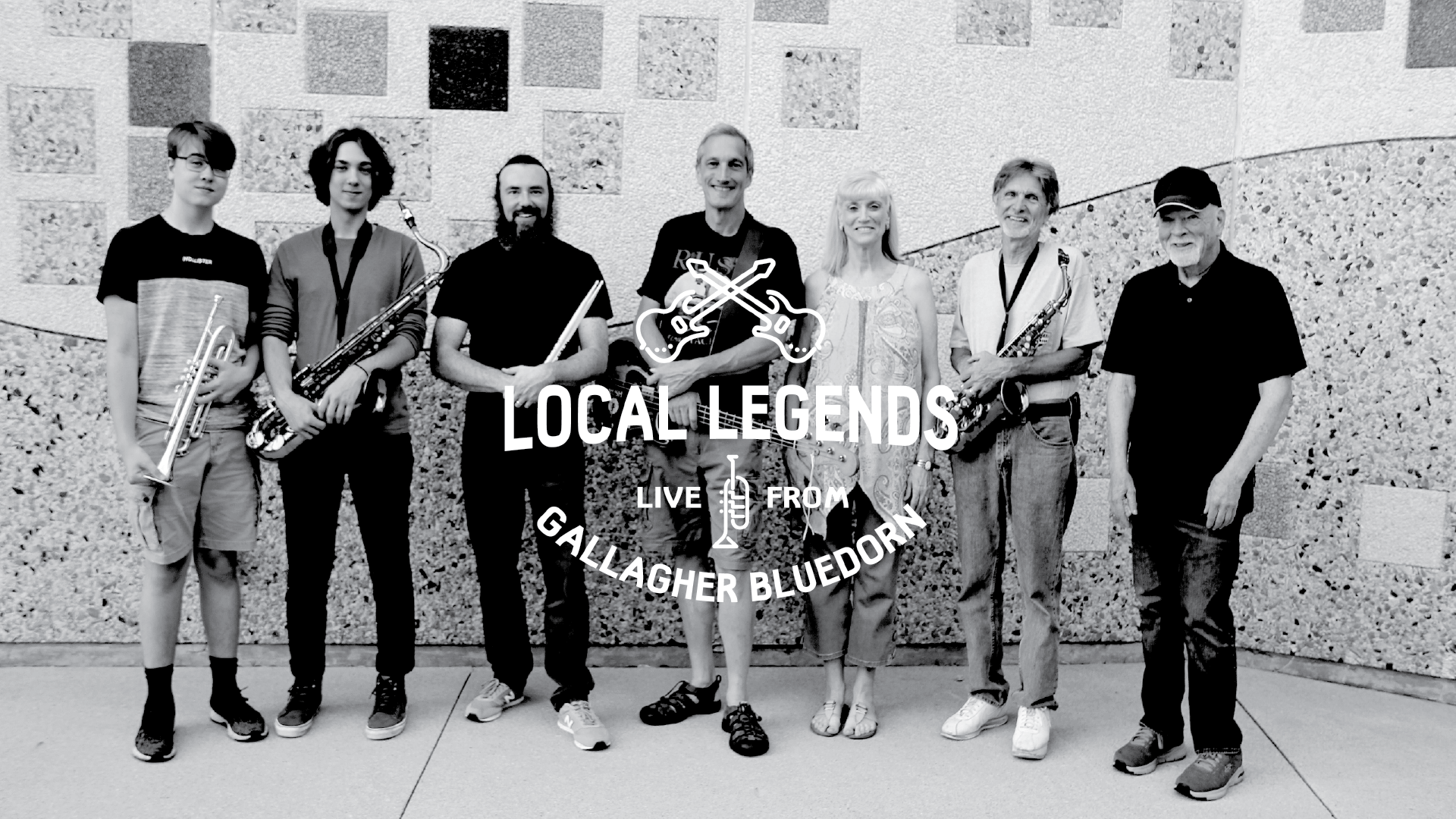 Seven members of Windsong stand against a brick wall and smile. The photo is in black and white with white Local Legends logo