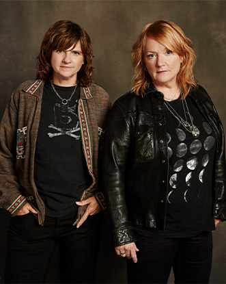 Two people dressed in black look at the camera with necklaces on. 