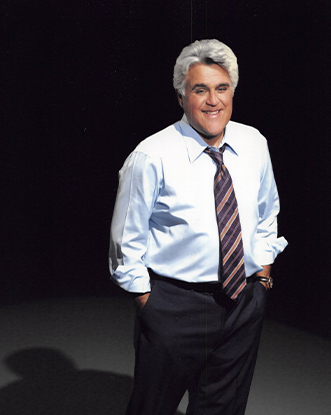 Jay Leno stands on an open stage with a white dress shirt and tie
