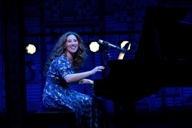 Actor playing Carole King sits at a piano and looks up smiling