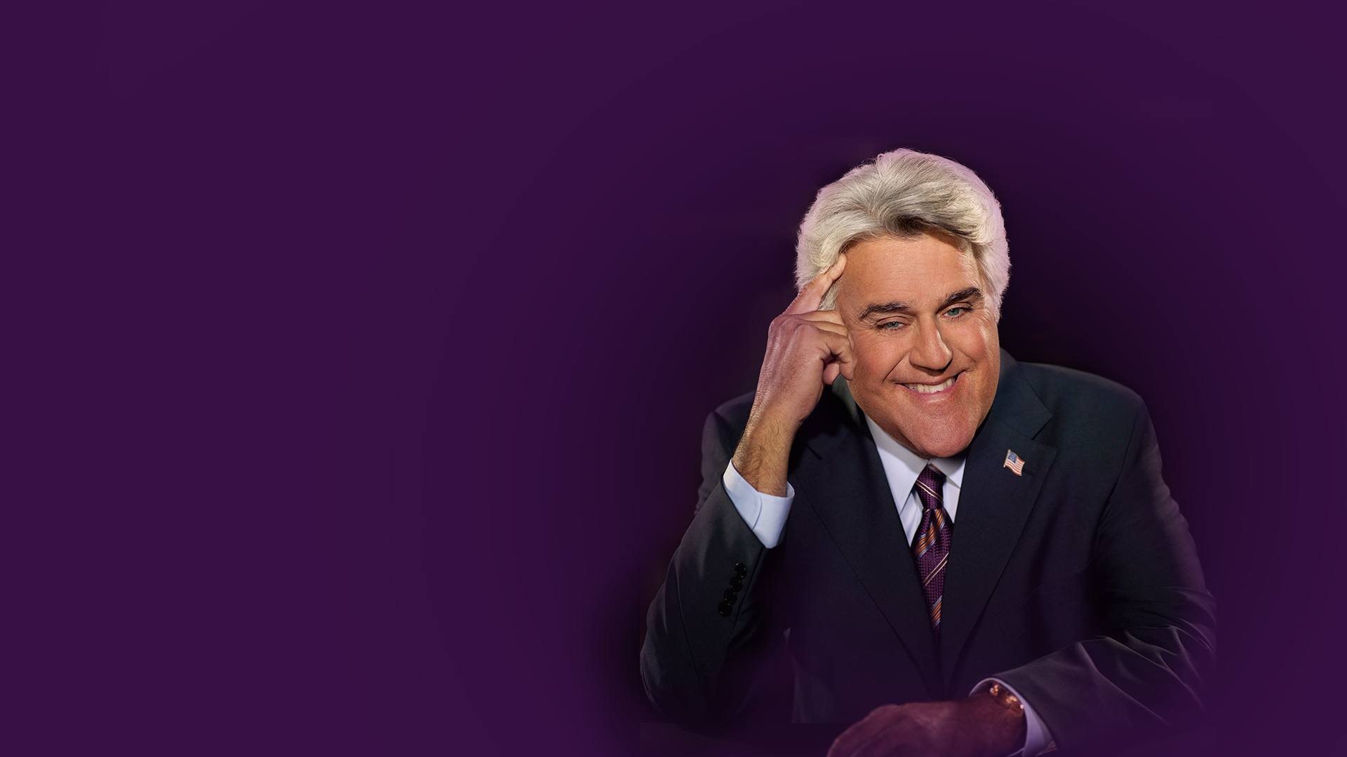 Jay Leno pointing to his temple and smiling. Background is deep purple