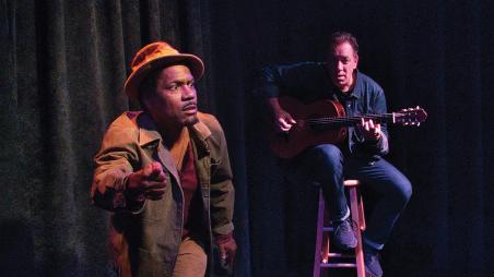 A man sitting down on a stool holding a guitar and a man with a brown hat running