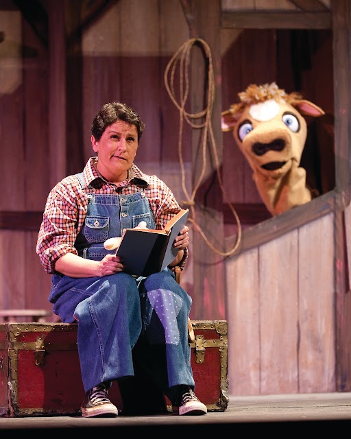 A person in overalls sitting down reading a book in a barn with stuffed animal cow looking in the background